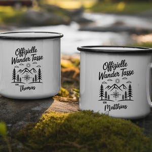 Puede incluir: Dos tazas de esmalte blanco con bordes y asas negros. Cada taza presenta un gr&aacute;fico negro de monta&ntilde;as, &aacute;rboles, una br&uacute;jula y el texto "Offizielle Wander Tasse" con un nombre debajo. Las tazas est&aacute;n al aire libre.