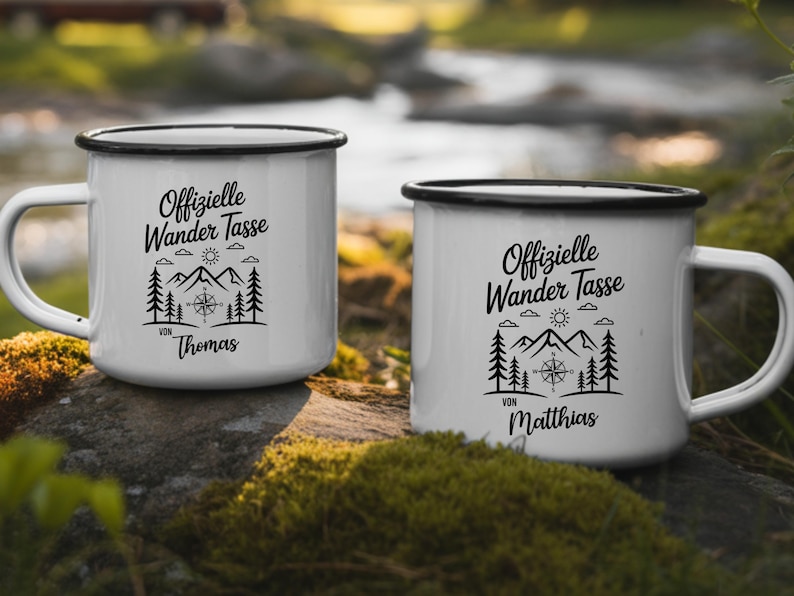Puede incluir: Dos tazas de esmalte blanco con bordes y asas negros. Cada taza presenta un gr&aacute;fico negro de monta&ntilde;as, &aacute;rboles, una br&uacute;jula y el texto "Offizielle Wander Tasse" con un nombre debajo. Las tazas est&aacute;n al aire libre.