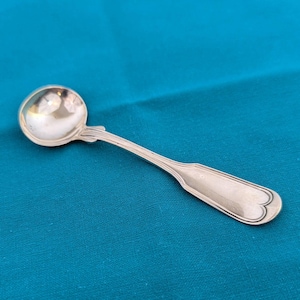 May include: A silver spoon with a round bowl and a long, ornate handle. The handle has a decorative design near the bowl and ends with a heart-shaped detail. The spoon is set against a teal background.