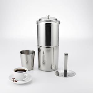 Puede incluir: Una cafetera de acero inoxidable con tapa, una taza y un platillo con café. La cafetera es cilíndrica y tiene un filtro y un soporte. La taza es blanca y está llena de café oscuro, con granos de café en el platillo.