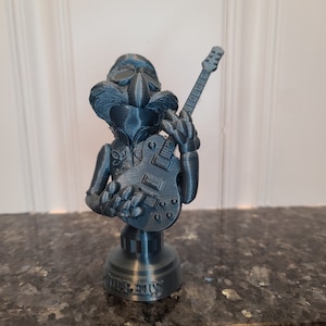 May include: A 3D printed figurine of a blue Muppet character holding a black electric guitar. The figurine is on a round base with the word "ROCKS" printed on it.
