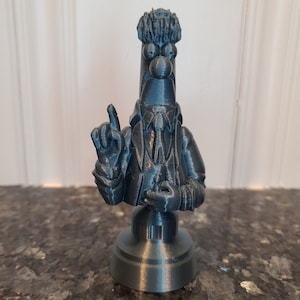 May include: A 3D printed bust of a cartoon character with a blue-gray finish. The character is wearing a suit and tie and has a surprised expression on its face. The bust is on a round base.