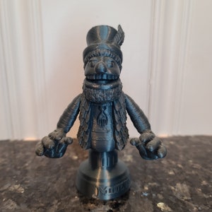 May include: A 3D printed figurine of a gray Muppet character wearing a black top hat and a feathered scarf. The figurine is standing on a gray base with the word "Muppet" printed on it.