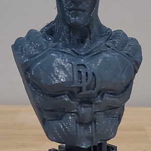 May include: A gray 3D printed bust of Daredevil, a superhero from Marvel Comics. The bust features a detailed face with horns and a classic Daredevil logo on the chest.