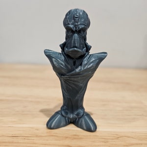 May include: A 3D printed gray bust of a creature with a large head and a detailed face. The creature is wearing a cloak that is draped over its shoulders and chest. The bust is standing on a wooden surface.