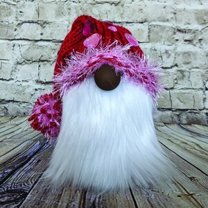 May include: A red knitted gnome hat with pink fur trim and pink heart embellishments. The gnome has a white fluffy beard.