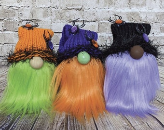 May include: Three Halloween gnomes with different coloured hair and hats. The gnome on the left has green hair and an orange hat with black trim. The gnome in the middle has orange hair and a purple hat with black trim. The gnome on the right has purple hair and a black hat with black trim.