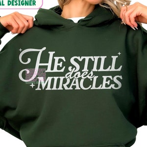 Może przedstawiać: Zielona bluza z kapturem z napisem "He Still Does Miracles" w białym kolorze. Bluzę nosi osoba siedząca.