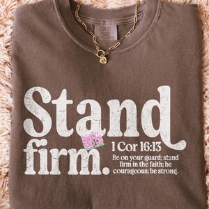 May include: A brown t-shirt with the words "Stand firm" in large white letters. Below the words is the Bible verse 1 Cor 16:13. A gold necklace with a pendant rests on the shirt. The shirt is on a fluffy, light brown surface.
