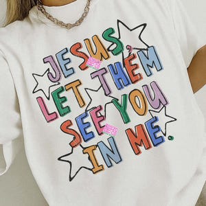 May include: White t-shirt with the text "JESUS LET THEM SEE YOU IN ME" in colorful, outlined letters. The design includes star shapes and the words "PNG SOLID & DISTRESSED". The shirt also has the text "2 DIFFERENT COLORS ORIGINAL DESIGNER".