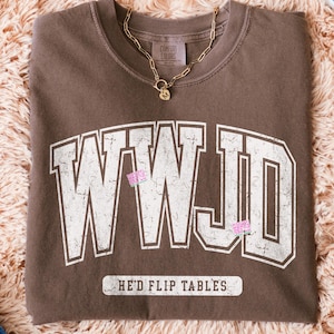 May include: A brown t-shirt featuring the text "WWJD" in a distressed white font. Below the text is "HE'D FLIP TABLES". A gold chain necklace with a pendant rests on the shirt. The shirt is on a fluffy, light brown surface.