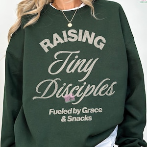 May include: Dark green sweatshirt with the text "RAISING Tiny Disciples" in a distressed, light gray font. Below the text, it reads "Fueled by Grace & Snacks". The sweatshirt has a crew neck and long sleeves.