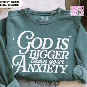 以下が含まれることがあります： ターコイズブルーのスウェットシャツに、「神はあなたの不安よりも大きい」という白い文字がかすれた感じで書かれています。テキストはヴィンテージスタイルのフォントです。