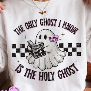 May include: A white crewneck sweatshirt featuring a graphic of a ghost holding a book labeled Holy Bible. The text reads The Only Ghost I Know Is The Holy Ghost. The design includes a black and white checkered pattern and small star accents.