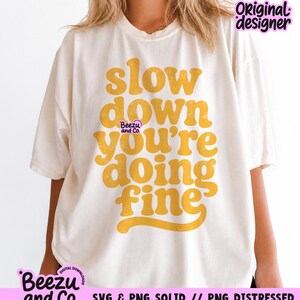May include: An off-white short-sleeve t-shirt featuring the mustard yellow text "slow down you're doing fine" in a bold, rounded font. A small "Beezu and Co." logo is printed in purple near the center, with "Original designer" in the top right.