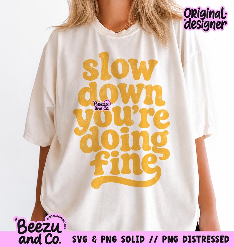 May include: An off-white short-sleeve t-shirt featuring the mustard yellow text "slow down you're doing fine" in a bold, rounded font. A small "Beezu and Co." logo is printed in purple near the center, with "Original designer" in the top right.