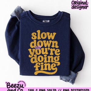 May include: A navy blue long-sleeve sweatshirt featuring the yellow text "slow down you're doing fine" in a distressed, rounded font. The garment has a crew neckline and ribbed cuffs, with a small "Beezu and Co." logo printed near the center text.