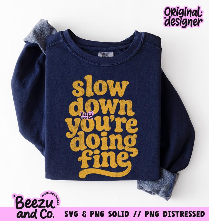 May include: A navy blue long-sleeve sweatshirt featuring the yellow text "slow down you're doing fine" in a distressed, rounded font. The garment has a crew neckline and ribbed cuffs, with a small "Beezu and Co." logo printed near the center text.