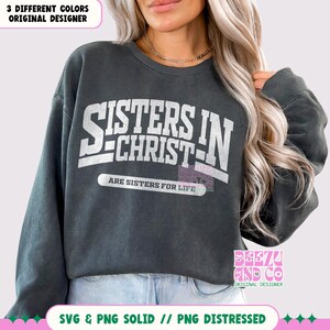 May include: A dark gray sweatshirt with the text "Sisters in Christ" in white lettering. The text is in a vintage style font and is surrounded by a white border. The text "Are Sisters for Life" is printed below the main text in a smaller font.