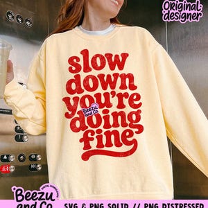 May include: A pale yellow crewneck sweatshirt featuring bold, distressed red text that reads "slow down you're doing fine" in a retro, rounded font. A small purple logo with the text "Beezu and Co." is centered within the graphic design.