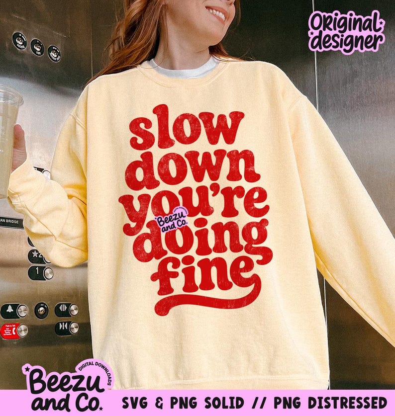 May include: A pale yellow crewneck sweatshirt featuring bold, distressed red text that reads "slow down you're doing fine" in a retro, rounded font. A small purple logo with the text "Beezu and Co." is centered within the graphic design.