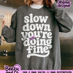 May include: A charcoal gray crewneck sweatshirt featuring white, distressed-style typography that reads, "slow down you're doing fine." The garment has long sleeves and a ribbed collar, with a small "Beezu and Co." logo printed in pink.