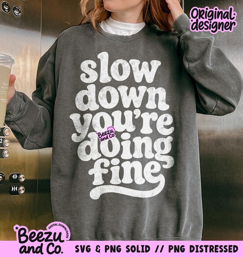May include: A charcoal gray crewneck sweatshirt featuring white, distressed-style typography that reads, "slow down you're doing fine." The garment has long sleeves and a ribbed collar, with a small "Beezu and Co." logo printed in pink.