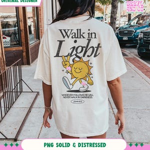 May include: Cream-colored t-shirt with the text "Walk in Light" and a cartoon sun character giving a peace sign. The shirt also features the Bible verse John 8:12. The shirt is a relaxed fit.