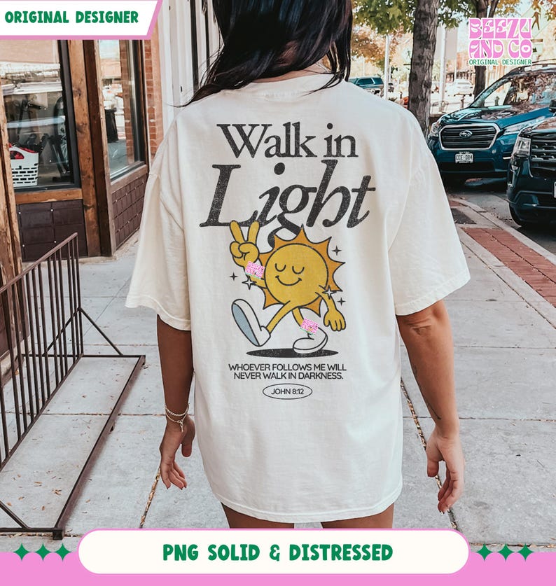 May include: Cream-colored t-shirt with the text "Walk in Light" and a cartoon sun character giving a peace sign. The shirt also features the Bible verse John 8:12. The shirt is a relaxed fit.