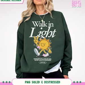 May include: Dark green sweatshirt with the text "Walk in Light" and a cartoon sun character giving a peace sign. The sweatshirt also features the text "WHOEVER FOLLOWS ME WILL NEVER WALK IN DARKNESS. JOHN 8:12".