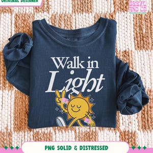 May include: A navy blue long-sleeve shirt with the words "Walk in Light" in white. The shirt features a smiling sun graphic with a peace sign and stars. The shirt is folded on a patterned surface.