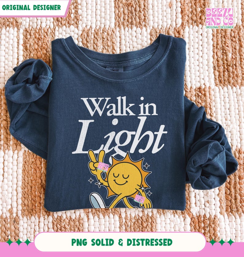 May include: A navy blue long-sleeve shirt with the words "Walk in Light" in white. The shirt features a smiling sun graphic with a peace sign and stars. The shirt is folded on a patterned surface.