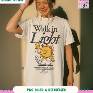 May include: White t-shirt with the words "Walk in Light" in black and a cartoon sun graphic. The sun is yellow with a smiling face and is making a peace sign. The text "WHOEVER FOLLOWS ME WILL NEVER WALK IN DARKNESS. JOHN 8:12" is below the graphic.