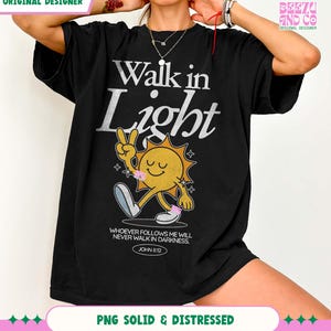 May include: Black t-shirt with the text "Walk in Light" in silver lettering. A cartoon sun character with a peace sign and the quote "Whoever follows me will never walk in darkness. John 8:12" is also featured.