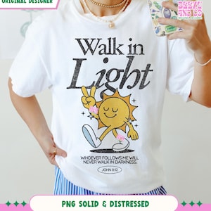 May include: White t-shirt with the text "Walk in Light" and a cartoon sun giving a peace sign. The sun has a smiling face and is wearing pink accessories. The shirt also features the Bible verse John 8:12.