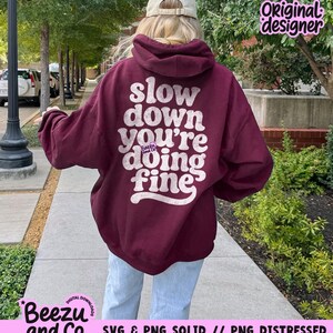 May include: A maroon hooded sweatshirt featuring a distressed white graphic on the back with the text "slow down you're doing fine" and a small "Beezu and Co." logo. The garment has long sleeves, a hood, and a relaxed fit, worn with light blue jeans.