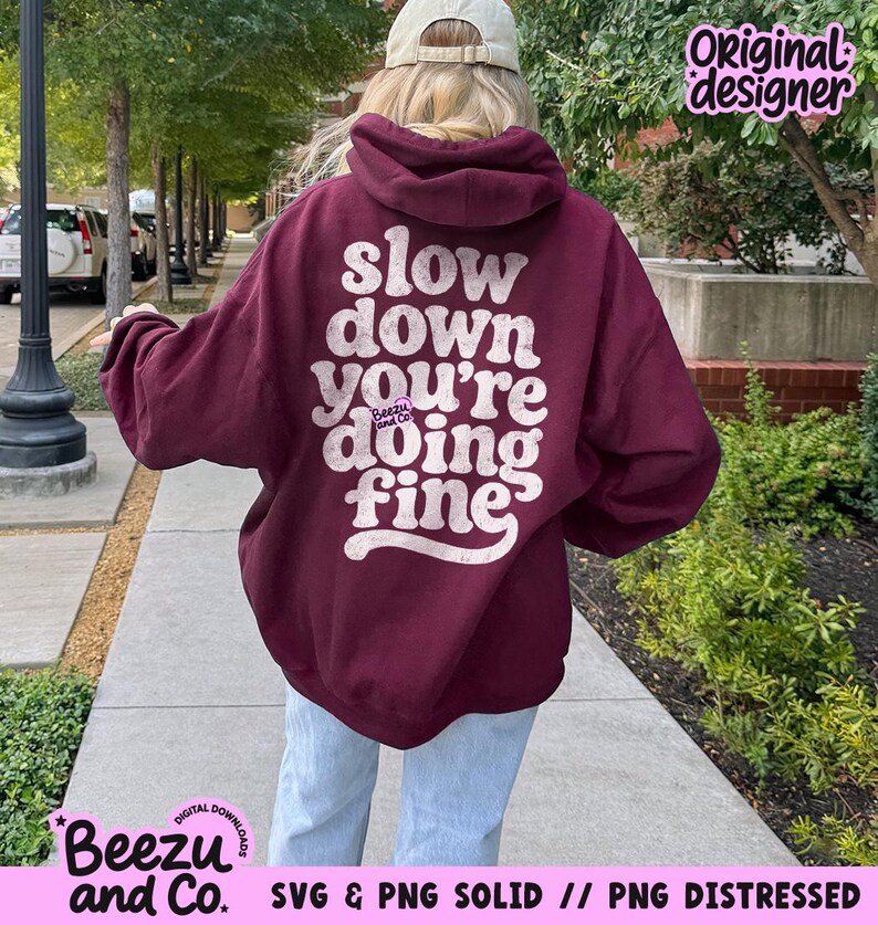 May include: A maroon hooded sweatshirt featuring a distressed white graphic on the back with the text "slow down you're doing fine" and a small "Beezu and Co." logo. The garment has long sleeves, a hood, and a relaxed fit, worn with light blue jeans.