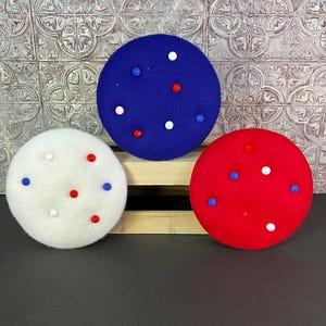 May include: Three round berets in red, white, and blue, each adorned with small, colorful, evenly spaced buttons. The berets are arranged on a wooden surface against a textured background, evoking a patriotic theme.