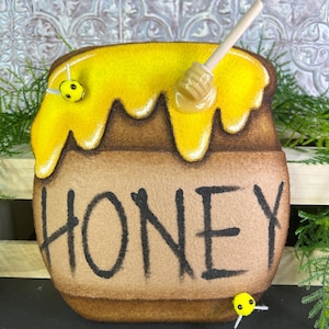 May include: A felt honey pot with yellow honey dripping from the top. The word "HONEY" is written in black on the front. A wooden honey dipper rests on top, and two small bee decorations are attached.