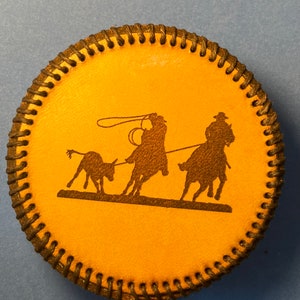 May include: Round, tan leather coaster with a black stitched edge. The center features a black silhouette of a cowboy roping a calf. A second cowboy on horseback is also in the image.