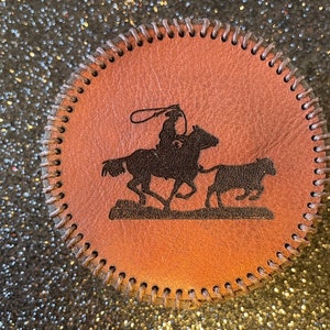 Pode incluir: Um descanso de copo de couro castanho com borda costurada apresenta uma silhueta de um cowboy a cavalo e a conduzir um bezerro.