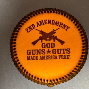 Könnte beinhalten: Ein brauner Lederuntersetzer mit einem schwarzen genähten Rand. Der Untersetzer zeigt eine Grafik mit zwei gekreuzten Gewehren mit dem Text "2nd Amendment", "God", "Guns* Guts" und "Made America Free!"