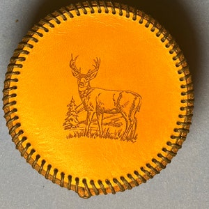 May include: Round, tan leather coaster with a stitched dark brown border. The center features a detailed engraving of a deer standing near a small tree. The coaster is a rustic, nature-themed item.