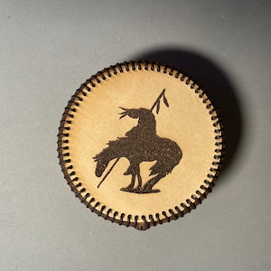 Puede incluir: Un posavasos redondo de cuero marrón con una silueta de un nativo americano montando un caballo. El posavasos tiene un borde cosido.