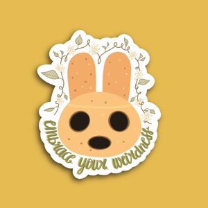 May include: A sticker featuring a cartoon animal with brown fur, large eyes, and a small mouth. The animal is surrounded by a wreath of green vines and white flowers. The text "Embrace your weirdness" is written in a cursive font around the animal.