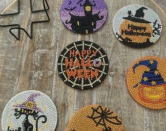 Op de afbeelding: Zes ronde, kleurrijke, glinsterende Halloween-onderzetters met verschillende ontwerpen, waaronder een spookhuis, een pompoen met een heksenhoed, een spinnenweb met de tekst "Happy Halloween", een pompoen met een heksenhoed, een zwarte kat met een heksenhoed en vleermuizen en een spinnenweb.