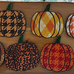 May include: Eight pumpkin-shaped diamond art coasters featuring various patterns including houndstooth, plaid, leopard print, and autumn leaves. Each coaster is decorated with small, colourful, reflective resin rhinestones in shades of orange, black, white, and green.