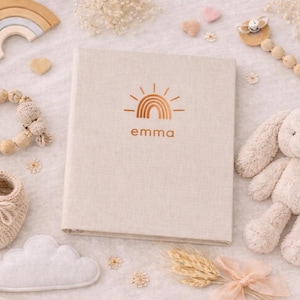 Könnte beinhalten: Ein beiges Baby-Erinnerungsbuch mit dem Namen "Emma" und einem Regenbogen-Design. Das Bild zeigt ein Plüschkaninchen, Holzspielzeug, Babyschuhe und eine wolkenförmige Dekoration, alles in sanften, neutralen Tönen.