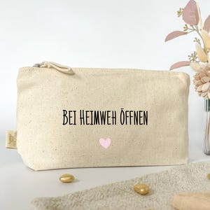 Könnte beinhalten: Eine beige Canvas-Kosmetiktasche mit Reißverschluss. Die Tasche hat schwarzen Text, der "Bei Heimweh Öffnen" lautet, und ein pinkfarbenes Herz.