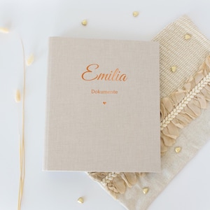 Könnte beinhalten: Ein beiges, mit Leinen bezogenes Buch mit dem Namen "Emilia" und dem Wort "Dokumente" in kupferfarbener Schrift. Unter dem Text befindet sich ein kleines kupferfarbenes Herz. Das Buch liegt auf einem beigen Dekostoff mit Rüschenkante.